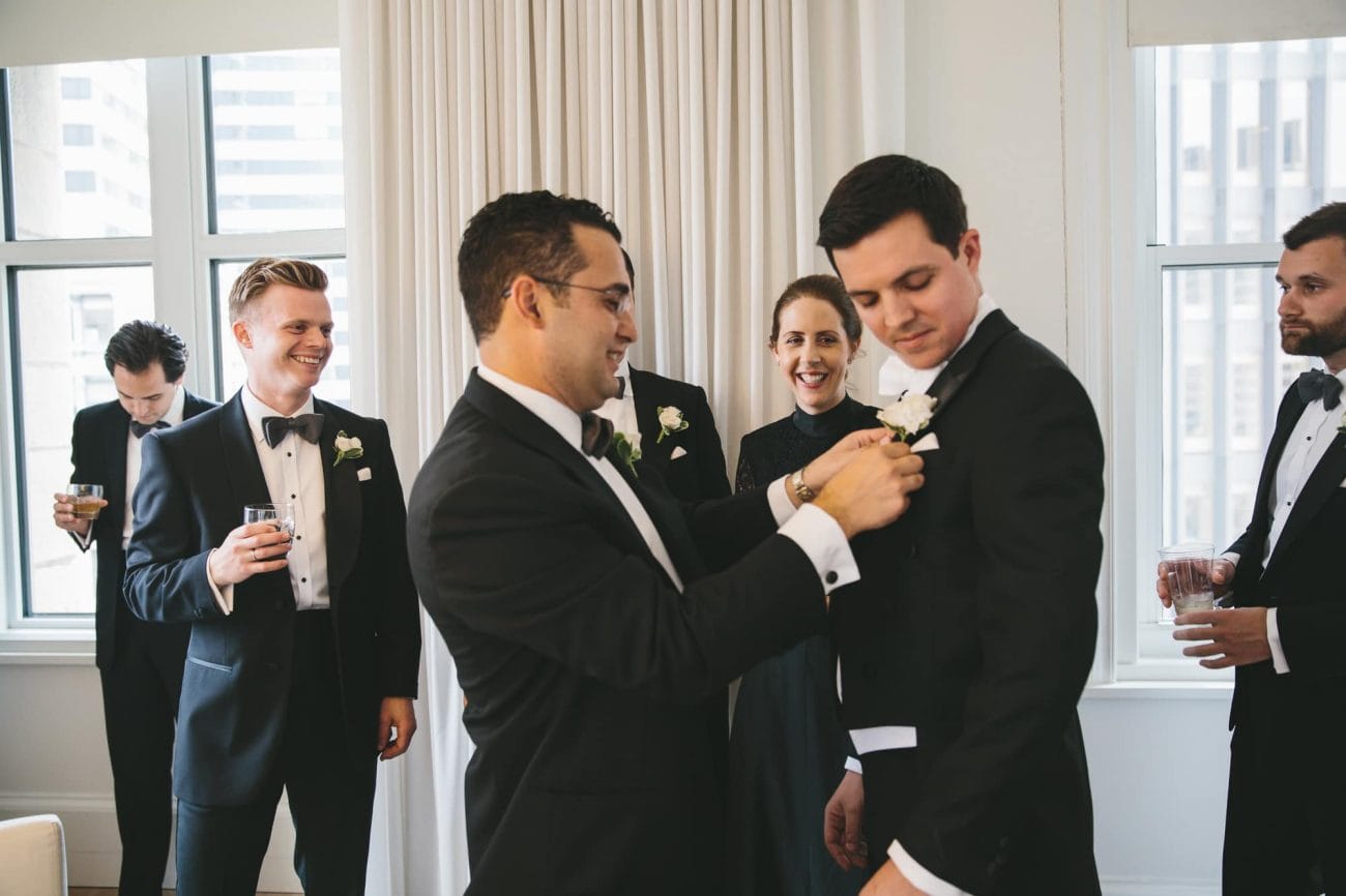 A documentary photograph of a groom getting his boutonnière on at the Ames Hotel before his State Room Wedding