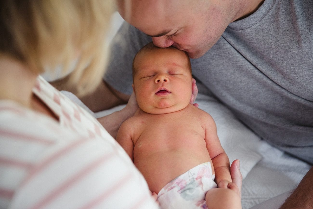 A documentary photograph of a father kissing his newborn daughter as they lay in bed during their lifestyle newborn session in Boston, Massachusetts