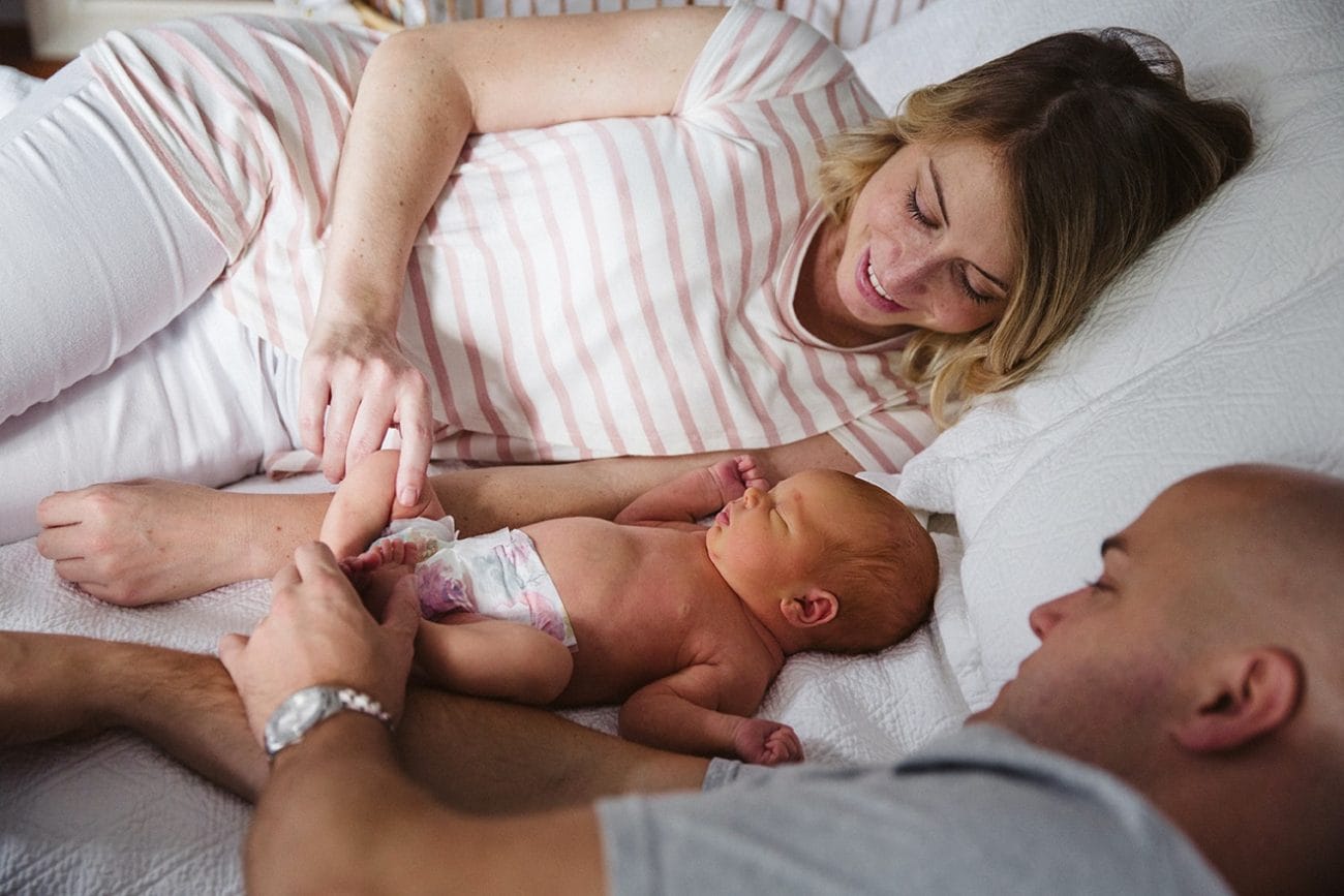 A documentary photograph of new parents laying in bed with newborn daughter during their lifestyle session at home in Boston, Massachusetts