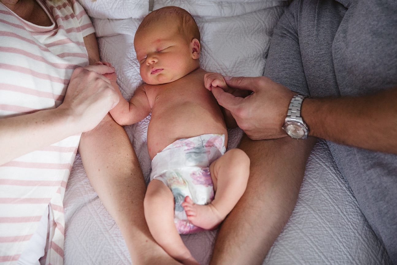 A documentary photograph of new parents holding their newborn daughter's hands as she lays in bed during their in home session in Boston, Massachusetts