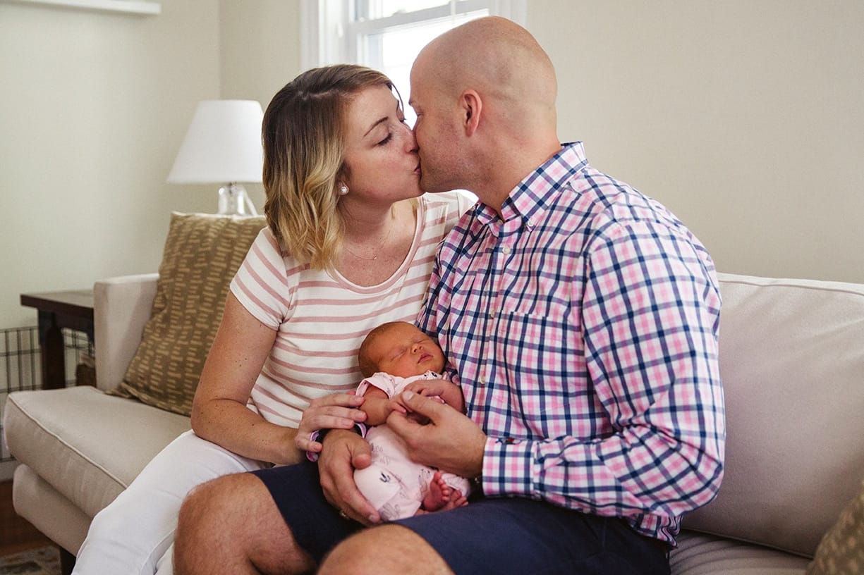 A documentary photograph of new parents kissing as they hold their newborn daughter during an in home lifestyle session in Boston, Massachusetts