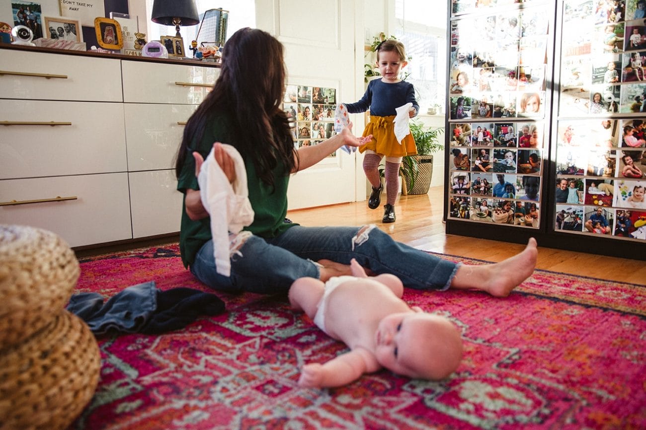 A documentary photograph of a little girl bringing her mom and baby brother a diaper and clothes during their in home family lifestyle session in Boston, Massachusetts