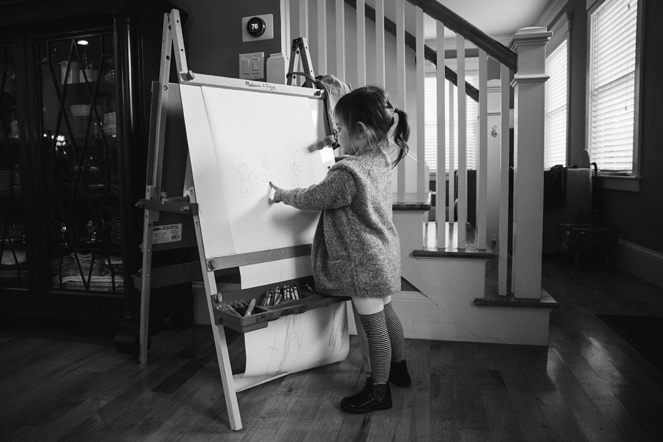 A documentary photograph of a little girl drawing at her easel during an in home family lifestyle session in Boston, Massachusetts