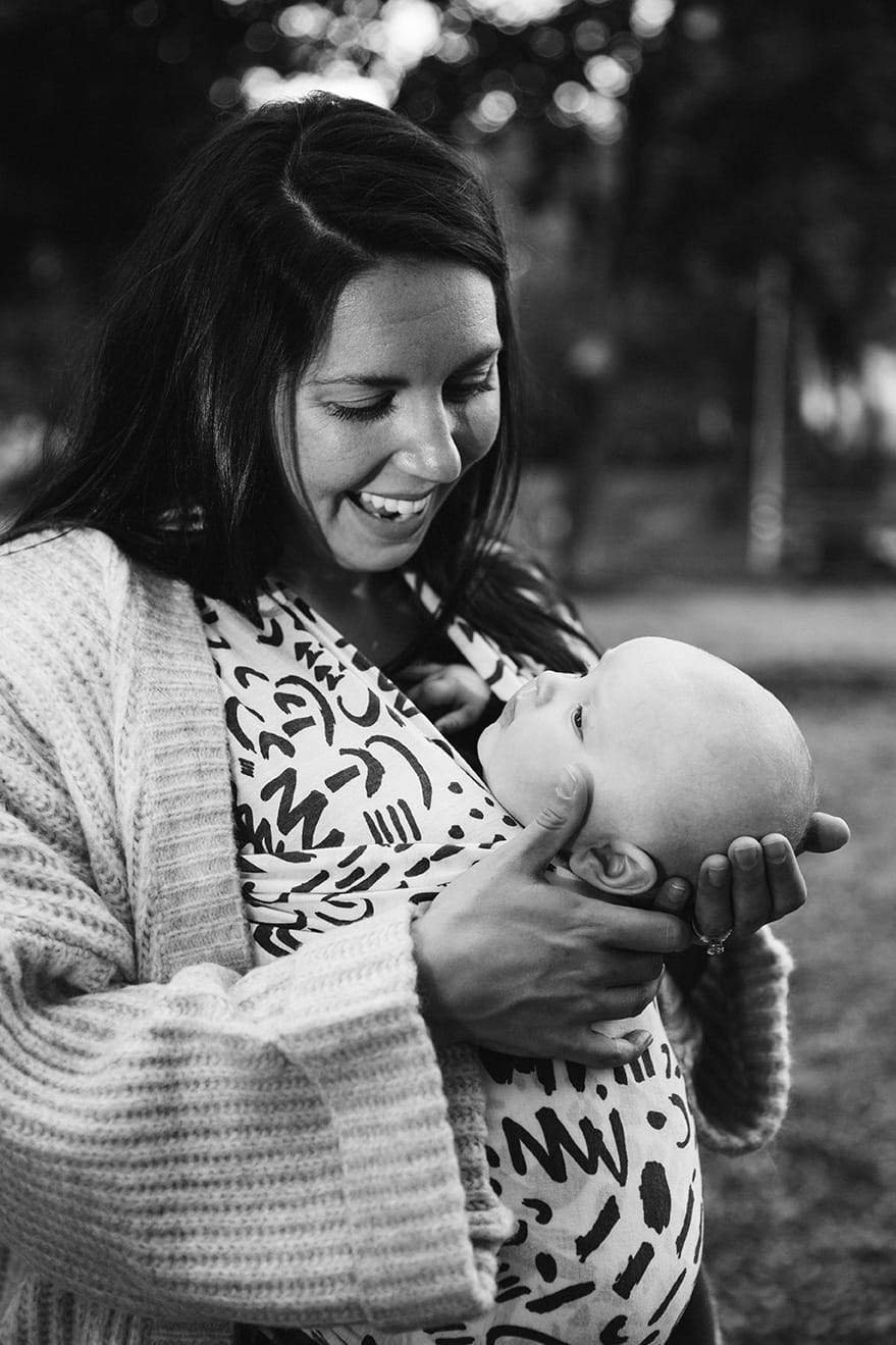 A documentary portrait of mom looking at her son while he's worn in a wrap at the playground during a family lifestyle session in Boston, Massachusetts