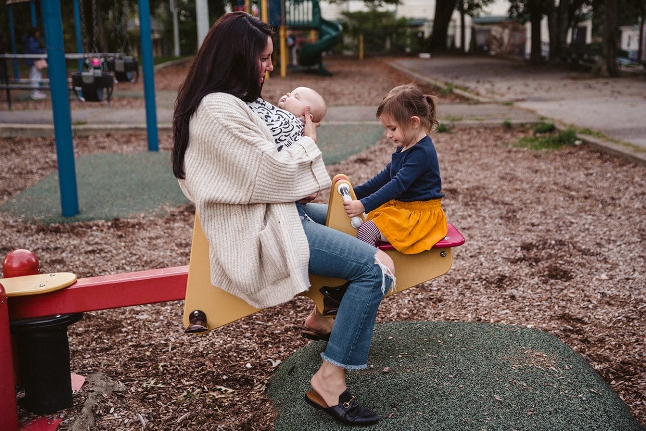A documentary photograph of a mom and baby bouncing on the see saw with the big sister during an in home family session in Boston, Massachusetts