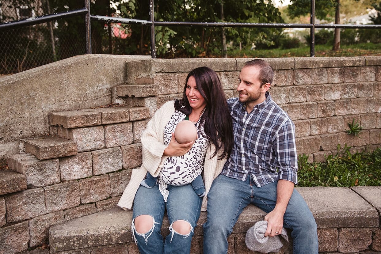 A documentary photograph of a mom and dad holding their baby boy and watching their daughter play at the playground during a family lifestyle session in Boston, Massachusetts