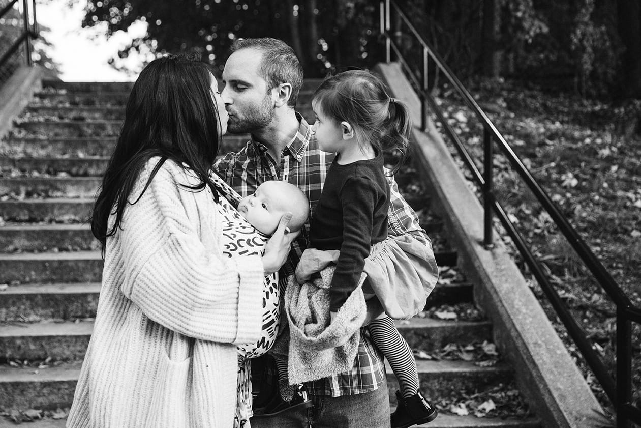 A documentary photograph of a husband and wife kissing while holding their kids during a family lifestyle session in Boston, Massachusetts