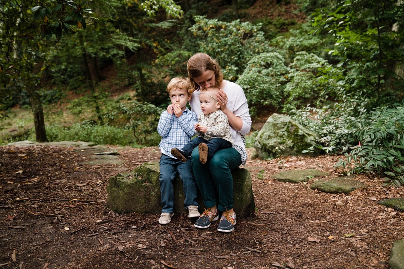 A documentary photograph of a mother cuddling and kissing her two kids as they have a snack during their family session at the Arnold Arboretum