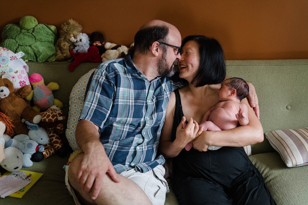 A documentary photograph of a couple kissing while holding their newborn daughter during their in home newborn session in Boston's Jamaica Plain