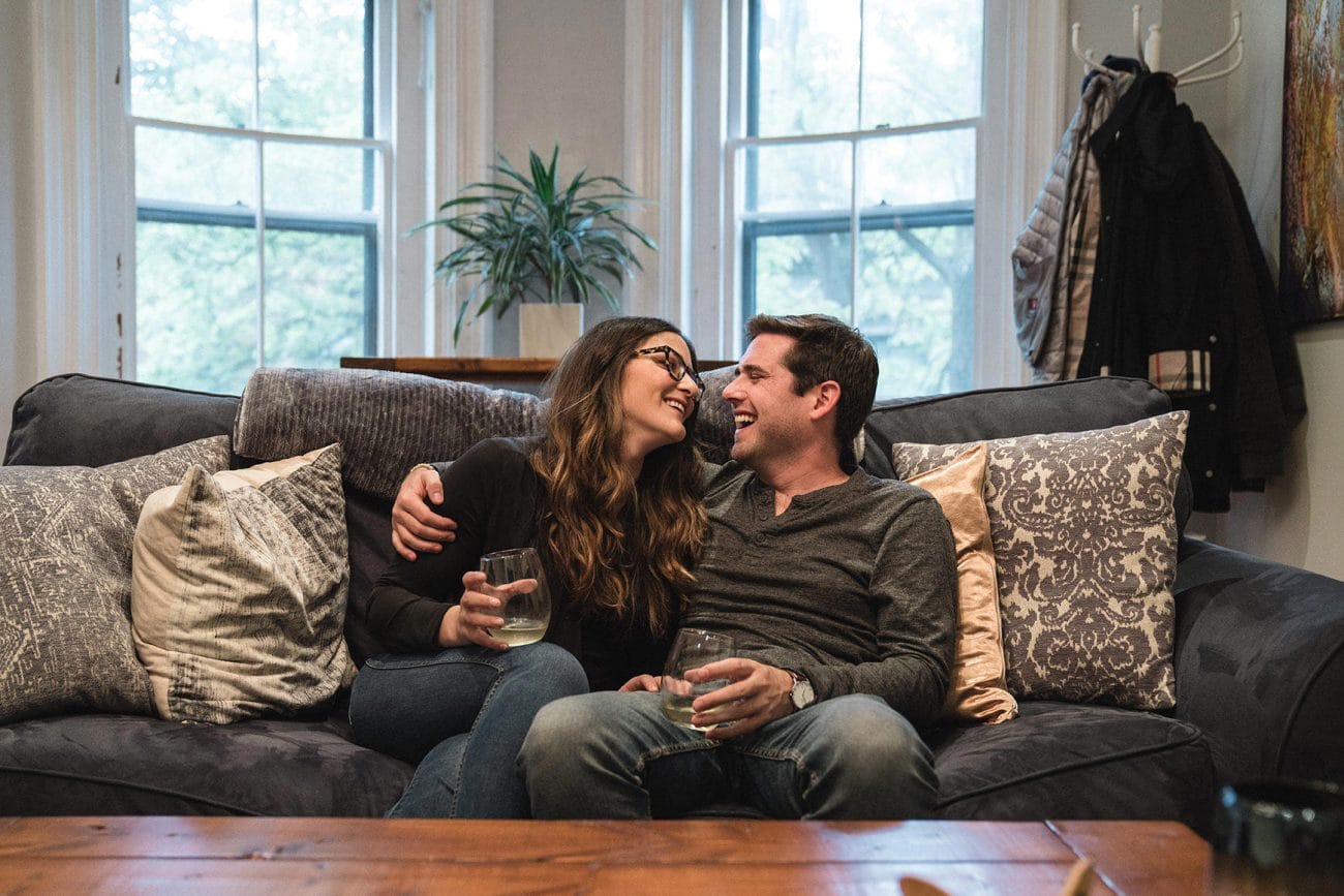 A documentary photograph of a couple laughing together on their couch during an in home boston engagement session