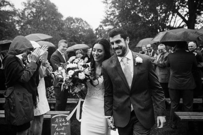 A documentary photograph of a couple walking up the aisle together after their outdoor ceremony in the rain