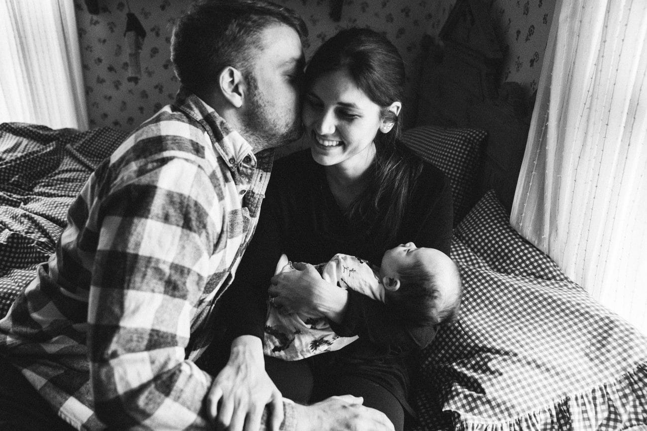 A documentary photograph of new parents giving each other a kiss while they hold their new baby during an in home family session in Boston