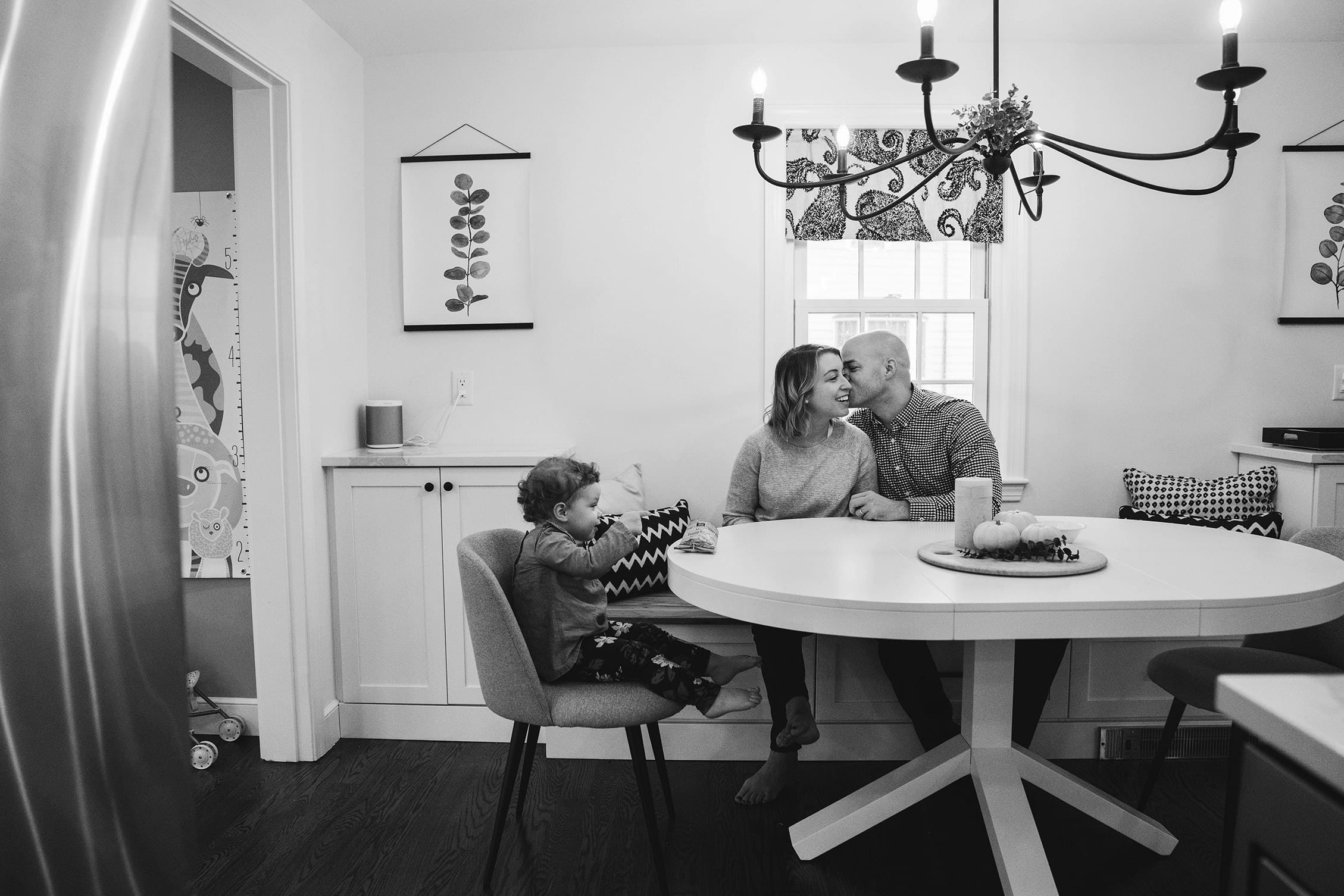 A documentary photograph of parents kissing while their daughter has a snack during an in home family session in Boston