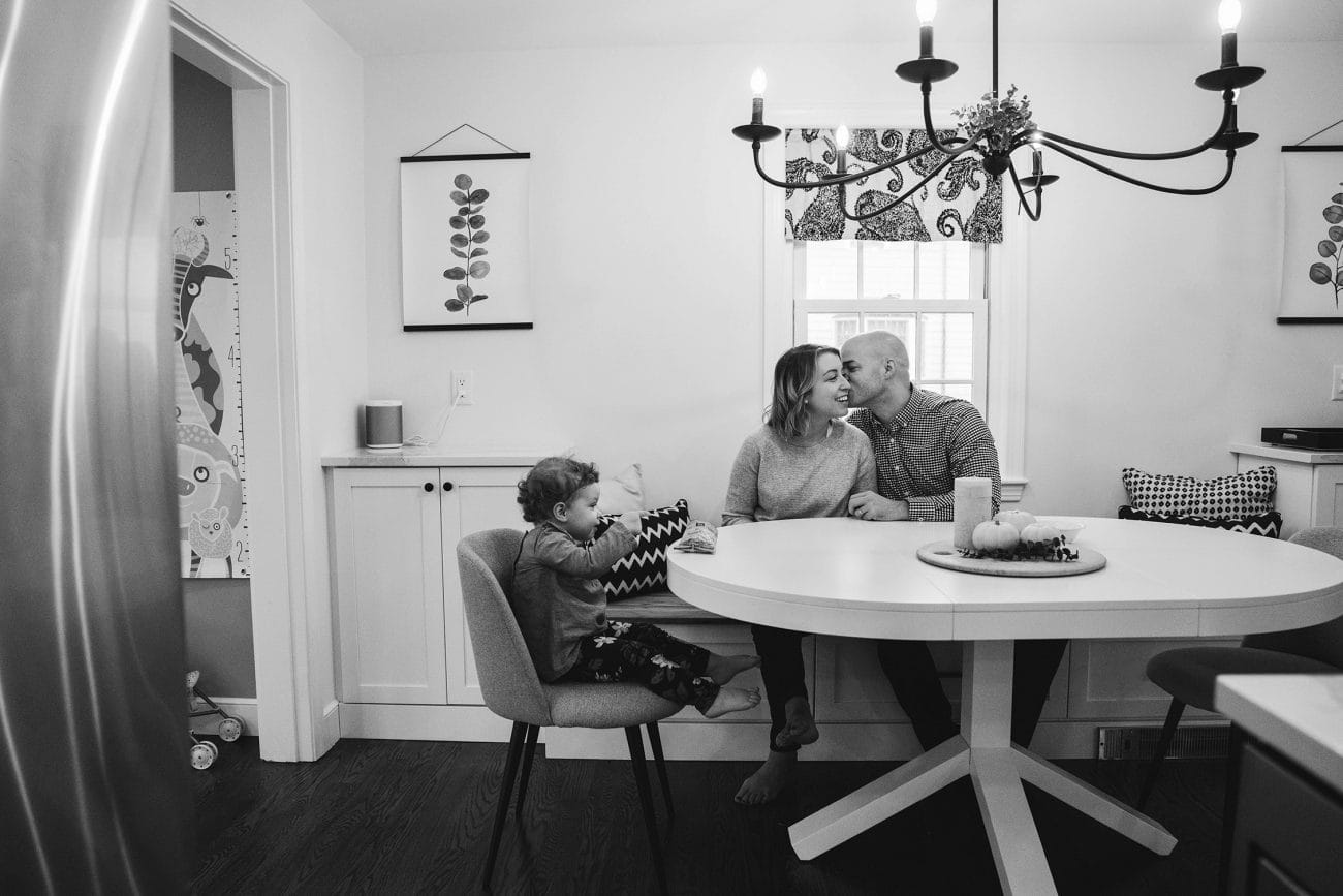 A documentary photograph of parents kissing while their daughter has a snack during an in home family session in Boston 