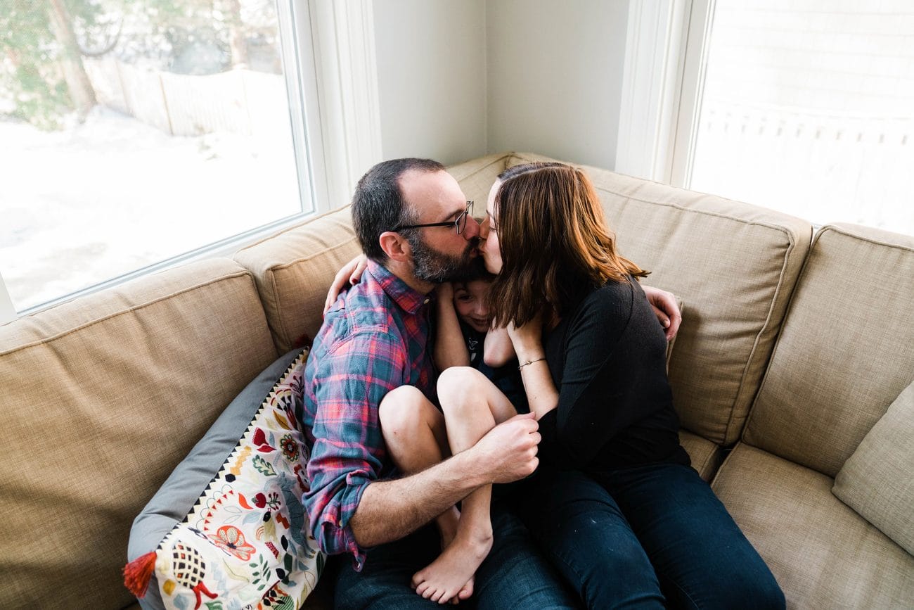 A documentary photograph of parents kissing and snuggling their son during an in home family session in Boston