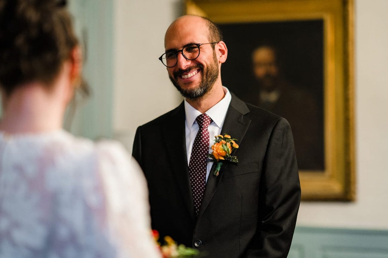 A documentary photograph of groom smiling at the bride during their Loring Greenough House Wedding ceremony.  