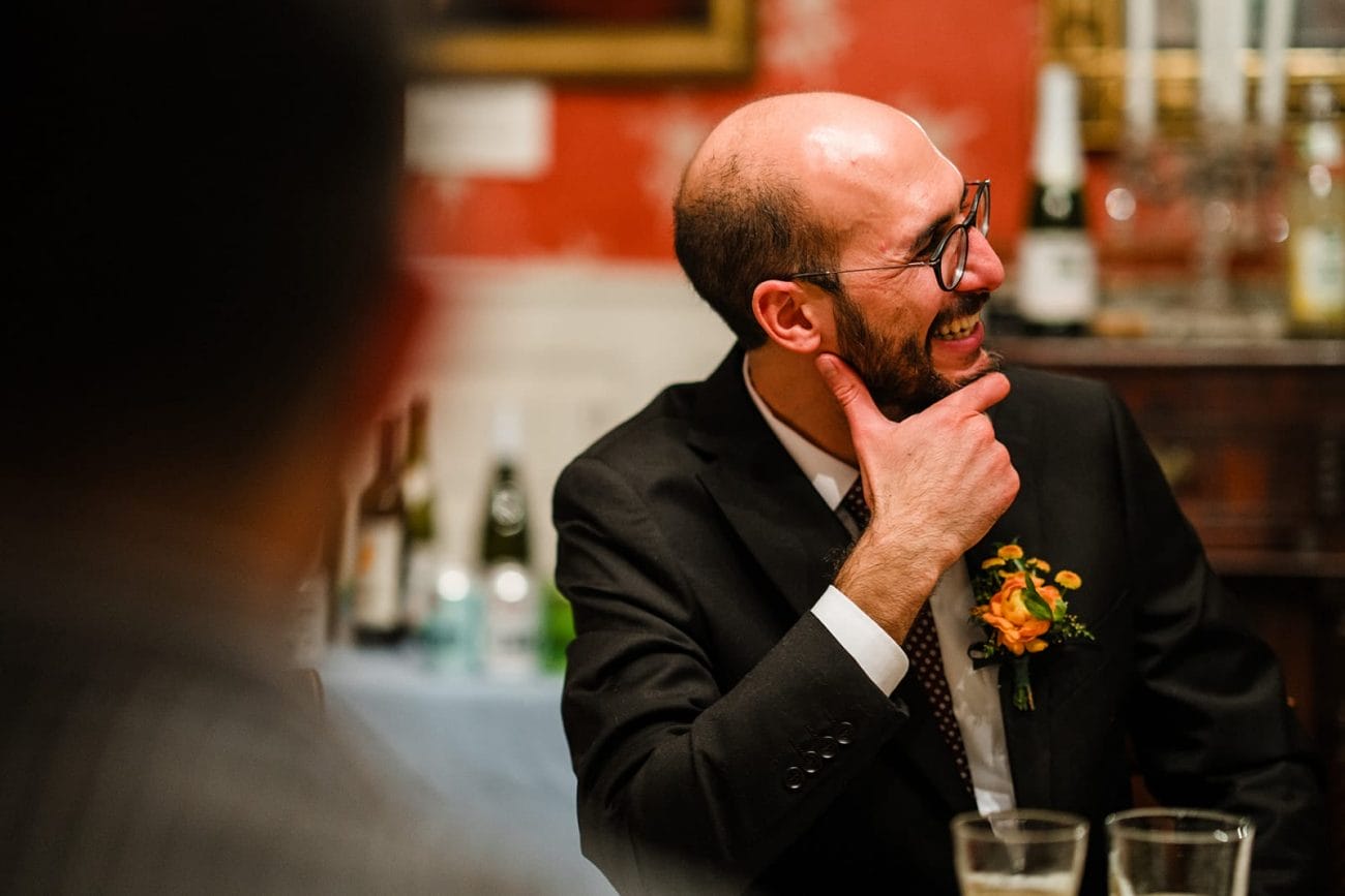 A documentary photograph of a groom talking and laughing with guests during the dinner portion of their Loring Greenough House Wedding.  